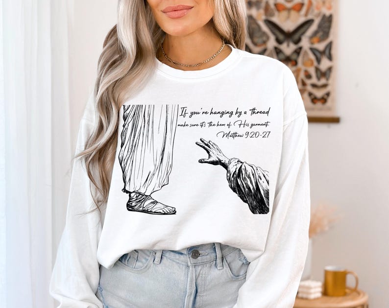 May include: White sweatshirt with a black and white illustration of a hand reaching out. The text on the sweatshirt reads "If you're hanging by a thread make sure it's the hem of His garment. Matthew 9:20-27".