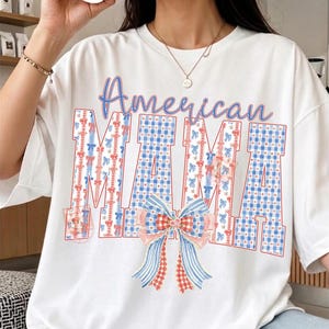 Op de afbeelding: Wit T-shirt met de tekst "American MAMA". Het woord "American" is in blauw geschreven, en "MAMA" is in een blauw-wit geruit patroon met rode en blauwe strikaccenten. Onderaan staat een grote strik.