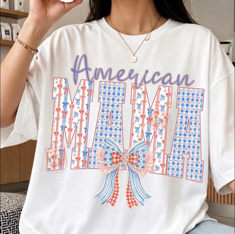 Op de afbeelding: Wit T-shirt met de tekst "American MAMA". Het woord "American" is in blauw geschreven, en "MAMA" is in een blauw-wit geruit patroon met rode en blauwe strikaccenten. Onderaan staat een grote strik.