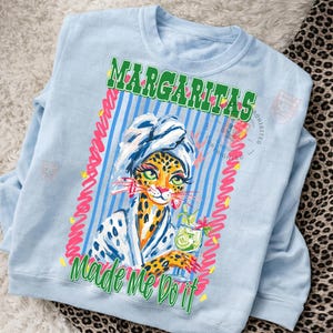 Pu&ograve; includere: Felpa azzurra con una grafica di un leopardo con un asciugamano e un accappatoio, che tiene in mano un cocktail. Il testo "Margaritas Made Me Do It" &egrave; stampato in verde e rosa.