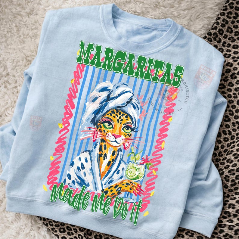 Pu&ograve; includere: Felpa azzurra con una grafica di un leopardo con un asciugamano e un accappatoio, che tiene in mano un cocktail. Il testo "Margaritas Made Me Do It" &egrave; stampato in verde e rosa.