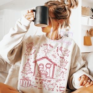 K&ouml;nnte beinhalten: Cremefarbenes Sweatshirt mit roter Grafik einer Krippenszene, eines Engels, eines Sterns und dem Text "Holy Night" und "The greatest gift of all". Ein gem&uuml;tliches Langarm-Oberteil.