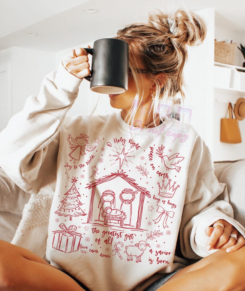 K&ouml;nnte beinhalten: Cremefarbenes Sweatshirt mit roter Grafik einer Krippenszene, eines Engels, eines Sterns und dem Text "Holy Night" und "The greatest gift of all". Ein gem&uuml;tliches Langarm-Oberteil.