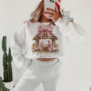 May include: White sweatshirt with a pink bow and a nativity scene graphic. The text "The greatest gift of all" and "Matthew 1:21" are printed below the image.
