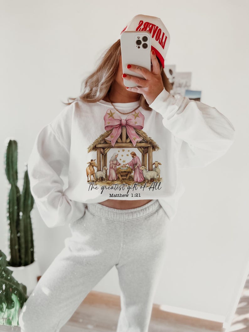 May include: White sweatshirt with a pink bow and a nativity scene graphic. The text "The greatest gift of all" and "Matthew 1:21" are printed below the image.