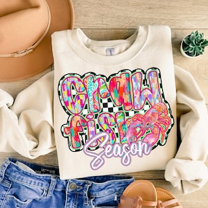 May include: A cream-colored sweatshirt with a colorful graphic design that says "Crawfish Season". The graphic features a pink crawfish with a checkered background.