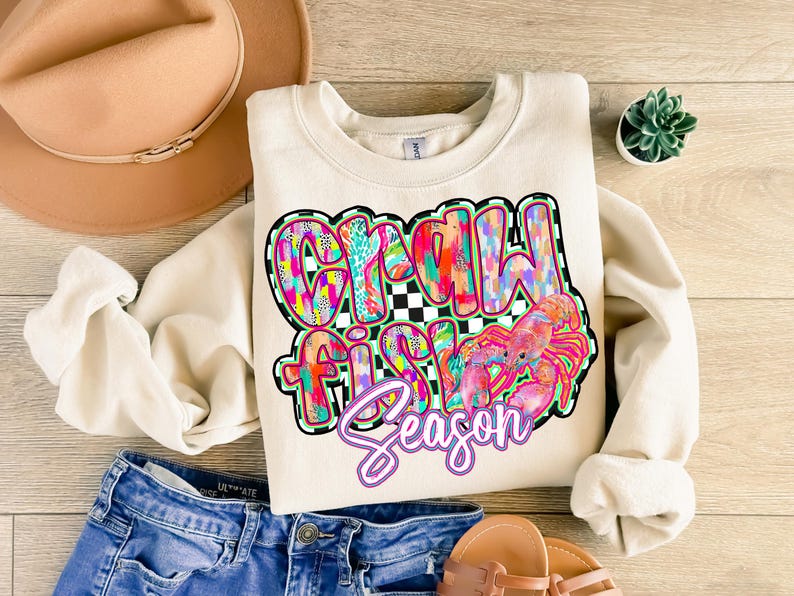 May include: A cream-colored sweatshirt with a colorful graphic design that says "Crawfish Season". The graphic features a pink crawfish with a checkered background.