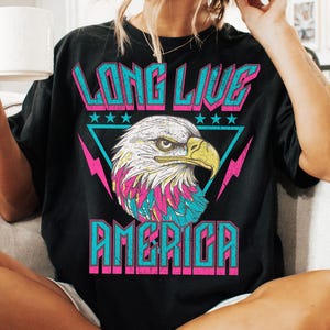 May include: Black t-shirt with a graphic print of an eagle with a pink, blue, and white design. The text "Long Live America" is printed in pink and blue letters.