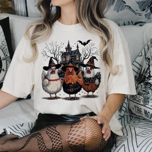 K&ouml;nnte beinhalten: Ein wei&szlig;es T-Shirt mit einem Aufdruck von drei H&uuml;hnern, die Hexenh&uuml;te tragen, vor einem Spukhaus. Die H&uuml;hner sind wei&szlig;, orange und schwarz-wei&szlig;.