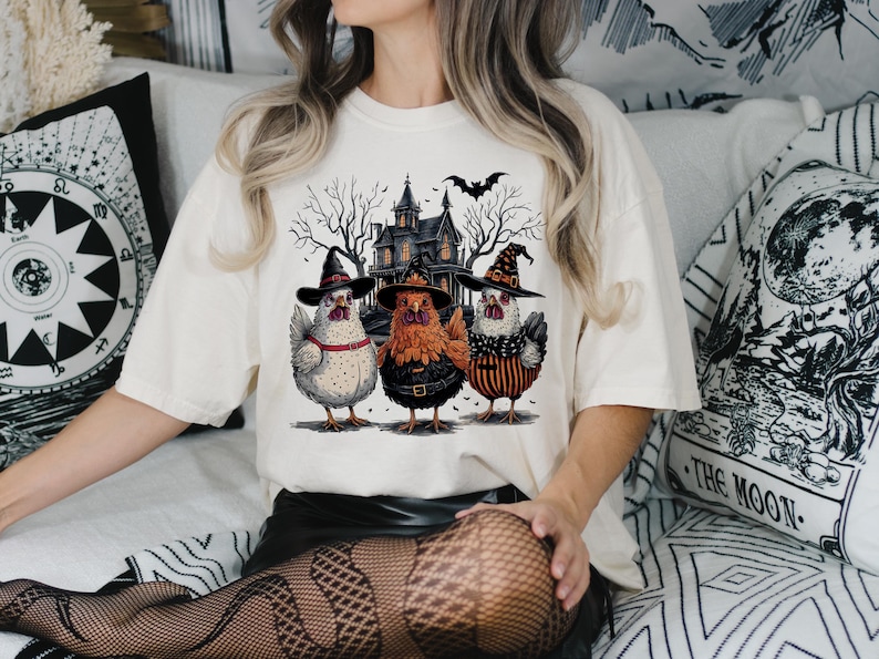 K&ouml;nnte beinhalten: Ein wei&szlig;es T-Shirt mit einem Aufdruck von drei H&uuml;hnern, die Hexenh&uuml;te tragen, vor einem Spukhaus. Die H&uuml;hner sind wei&szlig;, orange und schwarz-wei&szlig;.