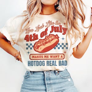 May include: Cream-colored t-shirt with a vintage-style graphic. The design features the text "You Look Like the 4th of July" above a hotdog illustration and the phrase "Makes Me Want a Hotdog Real Bad."