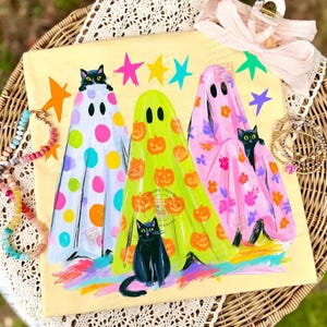 May include: A square cream-colored canvas featuring three ghosts in colorful sheets. The left ghost has multicolored polka dots, the center is lime green with orange pumpkins, and the right is pink with floral patterns. Black cats peek out.
