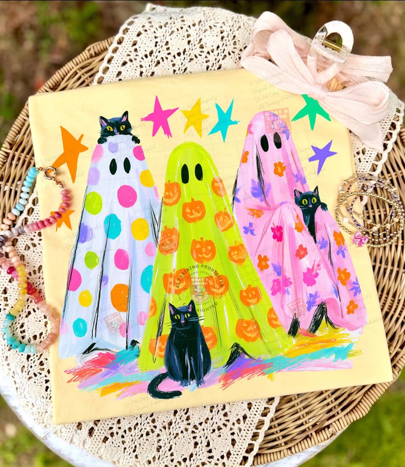 May include: A square cream-colored canvas featuring three ghosts in colorful sheets. The left ghost has multicolored polka dots, the center is lime green with orange pumpkins, and the right is pink with floral patterns. Black cats peek out.
