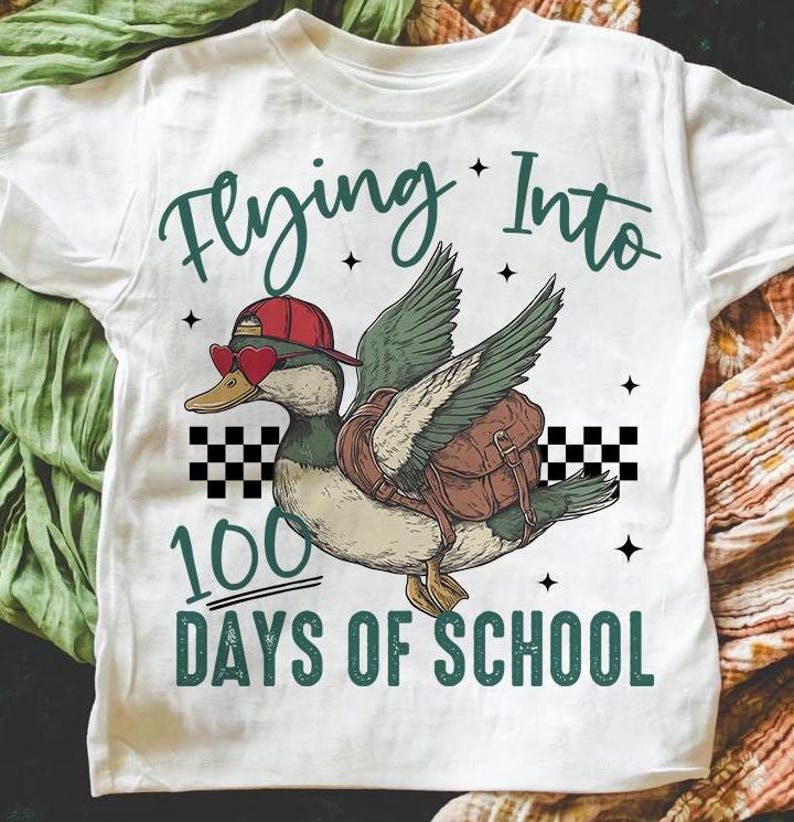 May include: White t-shirt featuring a cartoon duck wearing a red hat and sunglasses, flying with a backpack. The text "Flying Into 100 Days of School" is printed on the shirt.