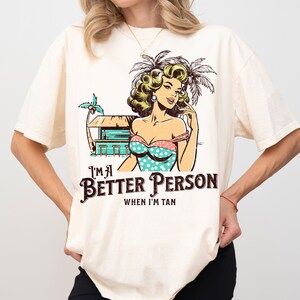 May include: A white t-shirt with a vintage-style illustration of a woman in a swimsuit and palm trees. The text "I'm a Better Person When I'm Tan" is printed on the shirt.