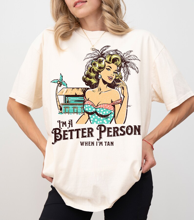 May include: A white t-shirt with a vintage-style illustration of a woman in a swimsuit and palm trees. The text "I'm a Better Person When I'm Tan" is printed on the shirt.