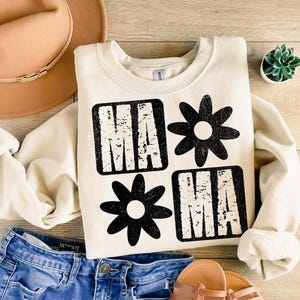 Peut inclure: Un sweat-shirt cr&egrave;me avec un motif graphique noir qui dit "MAMA" avec une marguerite entre les mots. Le graphique a un aspect vieilli.
