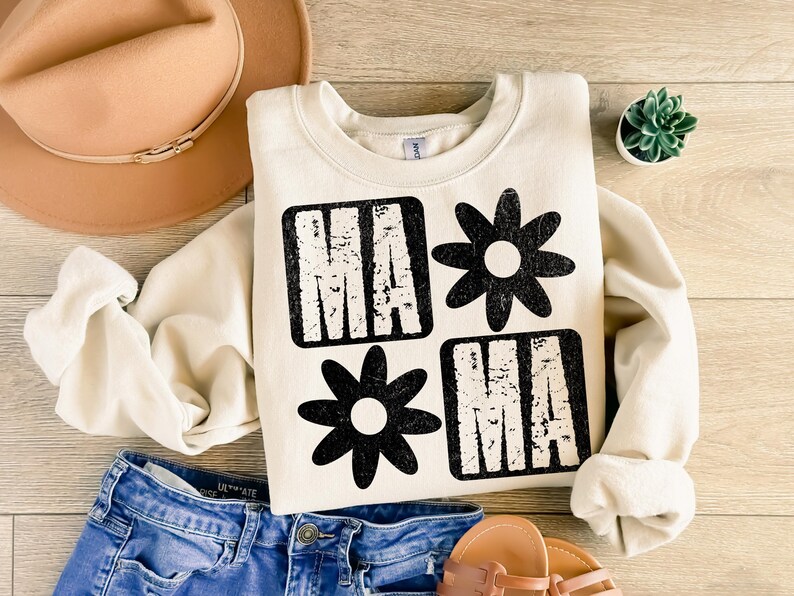 Peut inclure: Un sweat-shirt cr&egrave;me avec un motif graphique noir qui dit "MAMA" avec une marguerite entre les mots. Le graphique a un aspect vieilli.