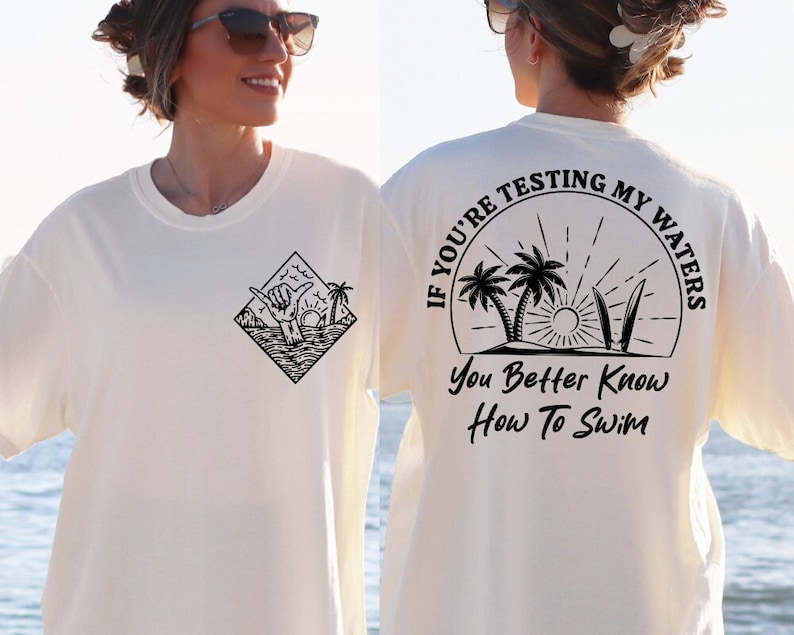 May include: White t-shirt with a black graphic of a hand giving the shaka sign with palm trees and waves. The back of the shirt has a black graphic circle with the text "If you're testing my waters you better know how to swim".