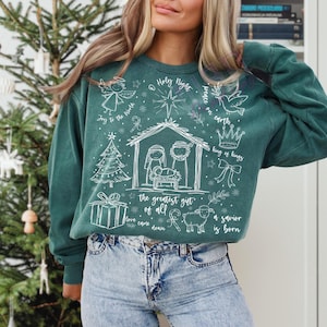 K&ouml;nnte beinhalten: T&uuml;rkisgr&uuml;nes Sweatshirt mit wei&szlig;en Illustrationen einer Krippenszene, eines Engels, eines Weihnachtsbaums und eines Geschenks. Der Text enth&auml;lt "Holy Night", "Joy to the World" und "A savior is born".