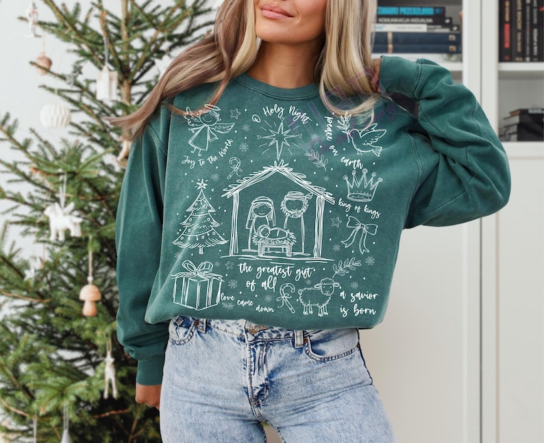 K&ouml;nnte beinhalten: T&uuml;rkisgr&uuml;nes Sweatshirt mit wei&szlig;en Illustrationen einer Krippenszene, eines Engels, eines Weihnachtsbaums und eines Geschenks. Der Text enth&auml;lt "Holy Night", "Joy to the World" und "A savior is born".