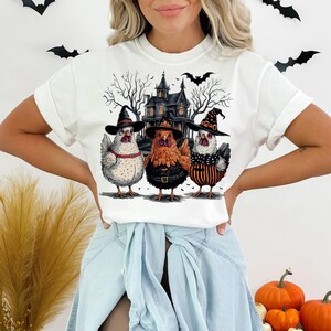K&ouml;nnte beinhalten: Wei&szlig;es T-Shirt mit Halloween-Motiv mit drei H&uuml;hnern, die Hexenh&uuml;te tragen, vor einem Spukhaus. Die H&uuml;hner sind wei&szlig;, orange und schwarz-wei&szlig;.