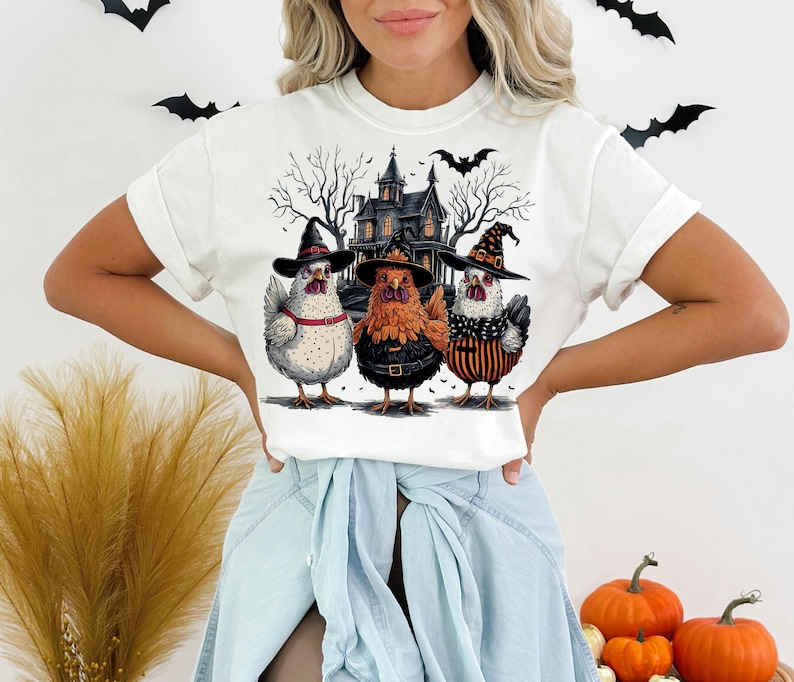 K&ouml;nnte beinhalten: Wei&szlig;es T-Shirt mit Halloween-Motiv mit drei H&uuml;hnern, die Hexenh&uuml;te tragen, vor einem Spukhaus. Die H&uuml;hner sind wei&szlig;, orange und schwarz-wei&szlig;.