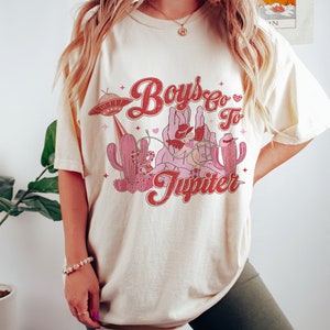 K&ouml;nnte beinhalten: Ein cremefarbenes T-Shirt mit einem Grafikdruck eines Cowboys, der auf einem Kaktus reitet, mit einem UFO im Hintergrund. Der Text auf dem Shirt lautet "Boys Go To Jupiter".