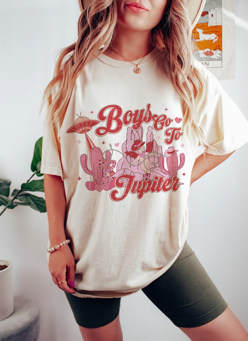K&ouml;nnte beinhalten: Ein cremefarbenes T-Shirt mit einem Grafikdruck eines Cowboys, der auf einem Kaktus reitet, mit einem UFO im Hintergrund. Der Text auf dem Shirt lautet "Boys Go To Jupiter".