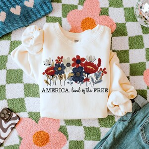 May include: A cream-colored sweatshirt with a floral design and the text "AMERICA. land of the FREE" printed on the front. The sweatshirt is lying on a green and white checkered surface.
