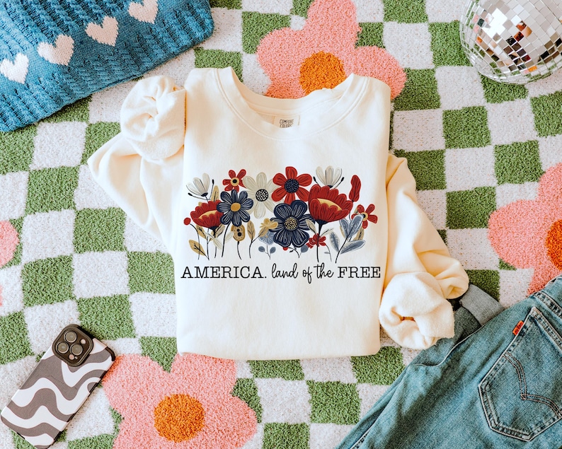 May include: A cream-colored sweatshirt with a floral design and the text "AMERICA. land of the FREE" printed on the front. The sweatshirt is lying on a green and white checkered surface.