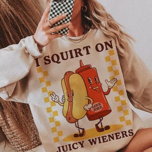 May include: A beige sweatshirt with a graphic of a hot dog and ketchup bottle with the text "I SQUIRT ON JUICY WIENERS" and "LOVE JUICE" printed on it.