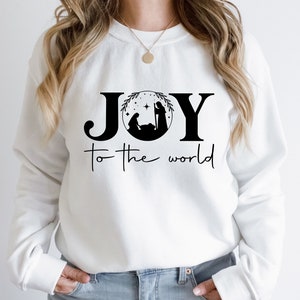 Pu&ograve; includere: Felpa bianca con un design grafico nero che dice "JOY to the world" con una scena della nativit&agrave; all'interno della "O".
