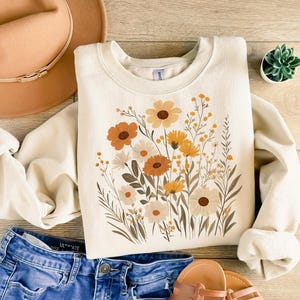 May include: A cream-colored sweatshirt with a floral print featuring yellow, orange, and brown flowers. The flowers are arranged in a bouquet-like design.