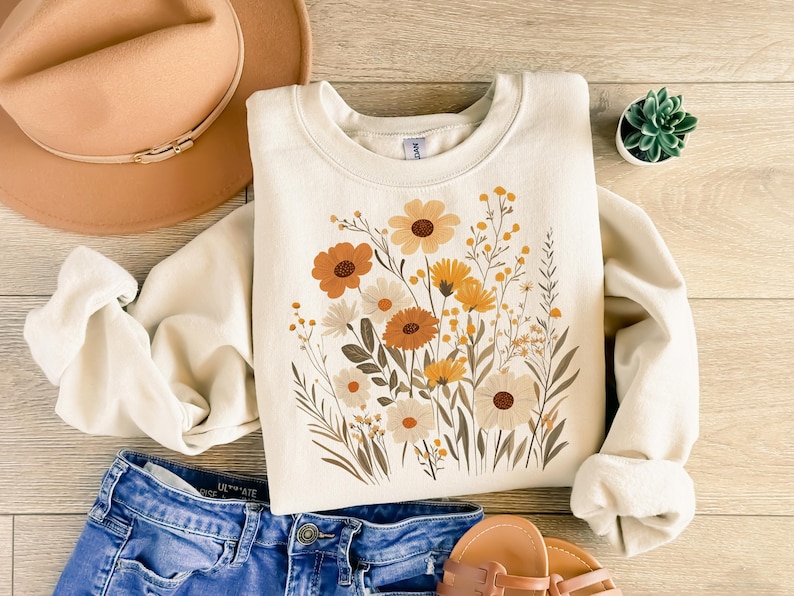 May include: A cream-colored sweatshirt with a floral print featuring yellow, orange, and brown flowers. The flowers are arranged in a bouquet-like design.
