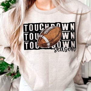 Op de afbeelding: Een beige sweatshirt met de tekst "TOUCHDOWN season" en een bruine en witte voetbalafbeelding.