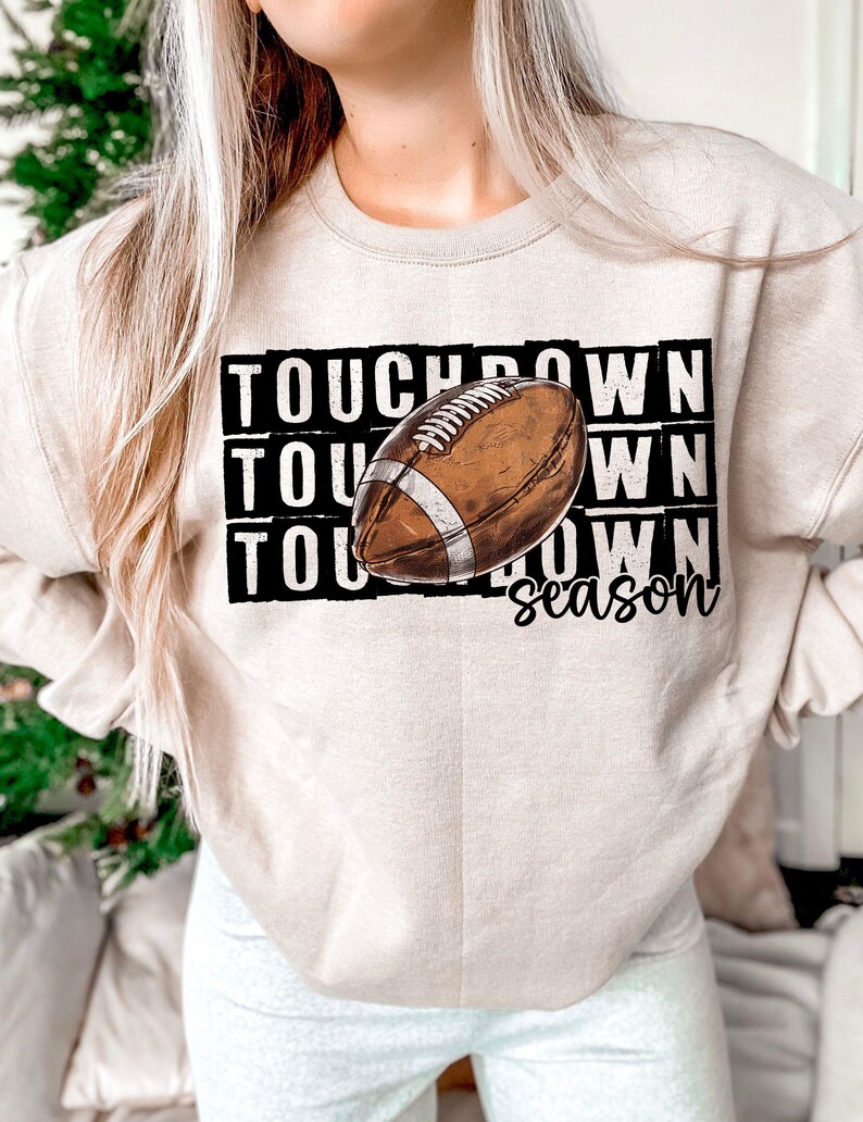 Op de afbeelding: Een beige sweatshirt met de tekst "TOUCHDOWN season" en een bruine en witte voetbalafbeelding.