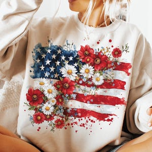 May include: A white sweatshirt with a watercolor design of an American flag with red, white, and blue flowers.