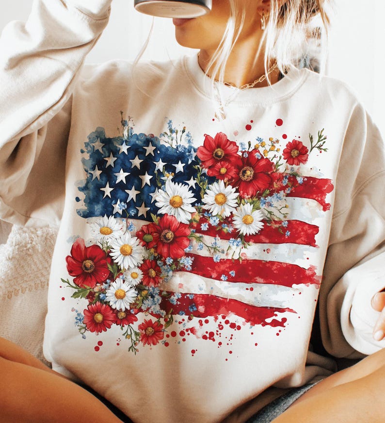 May include: A white sweatshirt with a watercolor design of an American flag with red, white, and blue flowers.