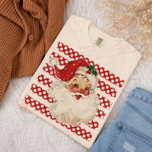 May include: A cream-colored t-shirt featuring a vintage-style illustration of Santa Claus with a red hat and white beard, centered over a background of horizontal red stripes with white polka dots. The shirt has a Comfort Colors brand label.