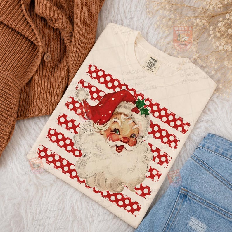 May include: A cream-colored t-shirt featuring a vintage-style illustration of Santa Claus with a red hat and white beard, centered over a background of horizontal red stripes with white polka dots. The shirt has a Comfort Colors brand label.