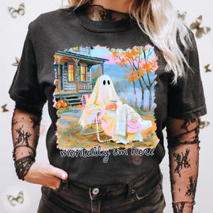 K&ouml;nnte beinhalten: Ein schwarzes T-Shirt mit einem Grafikdruck eines in ein wei&szlig;es Laken geh&uuml;llten Geistes, der in einem Gartenstuhl vor einem Haus mit orangefarbenen Herbstb&auml;umen sitzt und einen Kaffee h&auml;lt. Der Text unter dem Bild lautet, mentally i'm here.