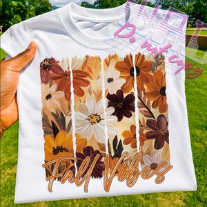 May include: White crew neck t-shirt featuring a floral design in shades of orange, brown, and white. The words "Fall Vibes" are printed in an orange script font at the bottom of the design. The shirt is held by a person.