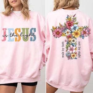 May include: A light pink sweatshirt with the text "Jesus is the light" in colorful letters on the front. The back features a floral cross design with the words "walk by faith, not by sight".