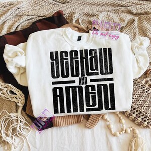May include: A white sweatshirt with the text "YEEHAW AND AMEN" in a bold, black, distressed font. The sweatshirt is folded on a bed with other clothing and accessories.