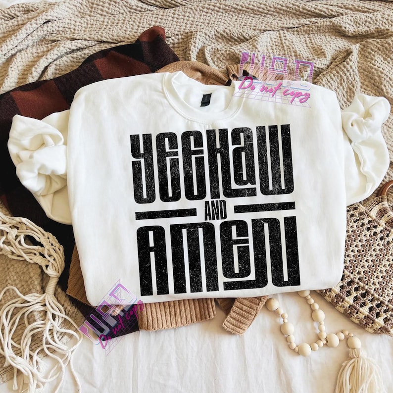 May include: A white sweatshirt with the text "YEEHAW AND AMEN" in a bold, black, distressed font. The sweatshirt is folded on a bed with other clothing and accessories.