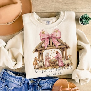 May include: A cream-colored sweatshirt with a pink bow and a nativity scene illustration. The text "The greatest gift of all" and "Matthew 1:21" are printed on the sweatshirt.