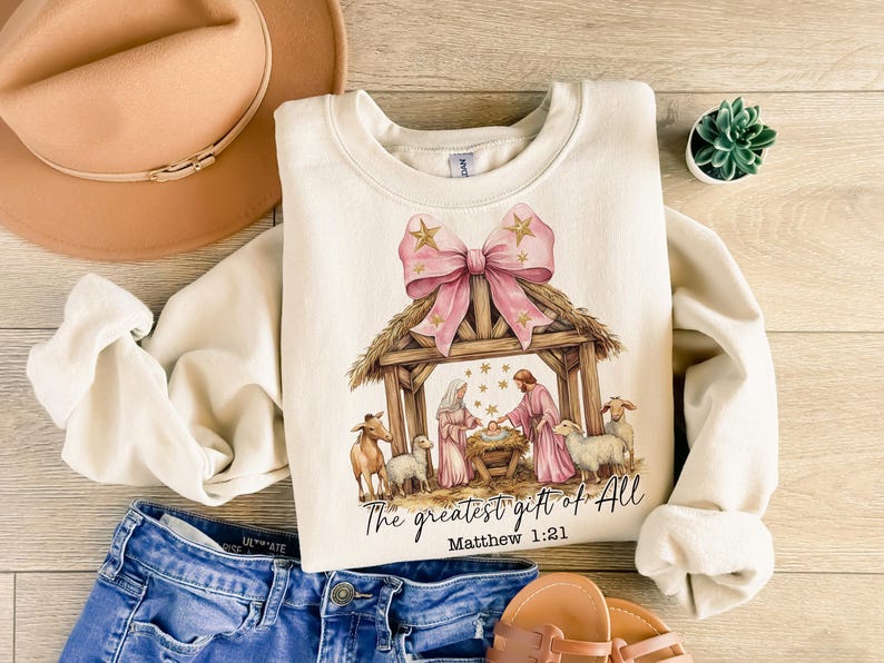 May include: A cream-colored sweatshirt with a pink bow and a nativity scene illustration. The text "The greatest gift of all" and "Matthew 1:21" are printed on the sweatshirt.