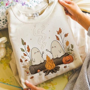 May include: Beige sweatshirt featuring a whimsical illustration of two friendly ghosts roasting marshmallows over a campfire. The design includes a pumpkin, autumn leaves, and a log. The sweatshirt is held up by a person.