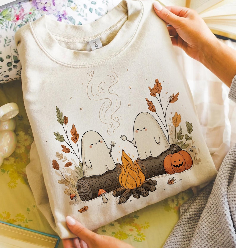 May include: Beige sweatshirt featuring a whimsical illustration of two friendly ghosts roasting marshmallows over a campfire. The design includes a pumpkin, autumn leaves, and a log. The sweatshirt is held up by a person.